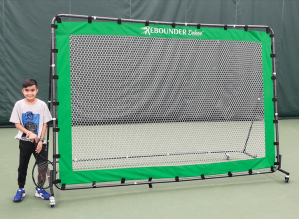 A young boy holds a tennis racket next to the rebounder deluxe tennis rebound net from oncourt offcourt. 